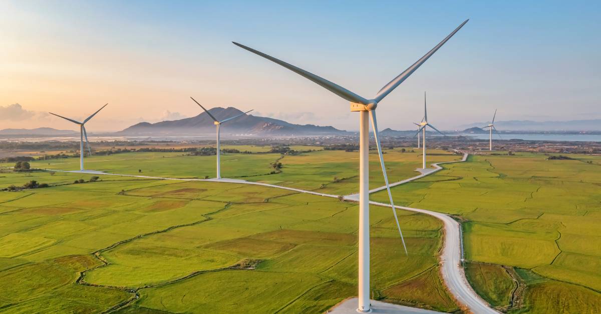 A panoramic view of a large field with mountains and a body of water in the distance. Several wind turbines are in the field.