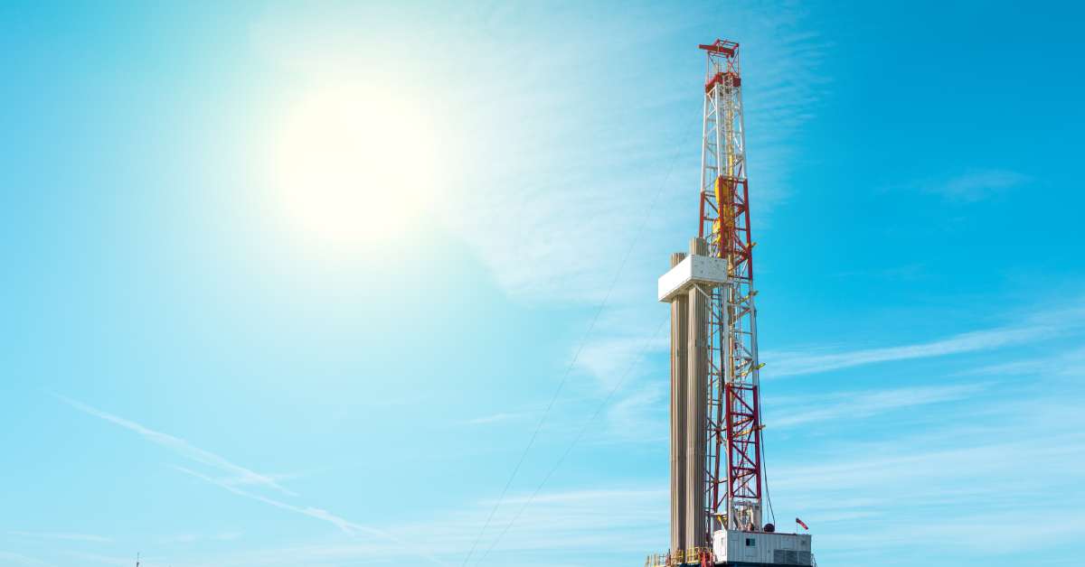A tall white and red land rig stands on a platform for oil drilling, surrounded by tall grass and a blue sky.